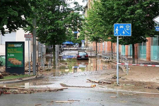 Teilweise überflutete kleine Straße, ein Auto steht im Wasser, Äste und Kleinholz liegen herum.