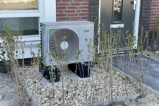 Eine Wärmepumpe, die wie eine Klimaanlage aussieht, steht auf Füßen in einem Steinbeet neben dem Eingang eines Einfamilienhauses.