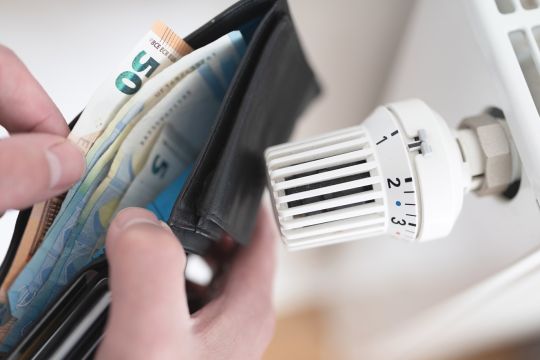 Hände halten eine Geldbörse mit einigen Euro-Scheinen, daneben der Thermostat eines Heizkörpers.